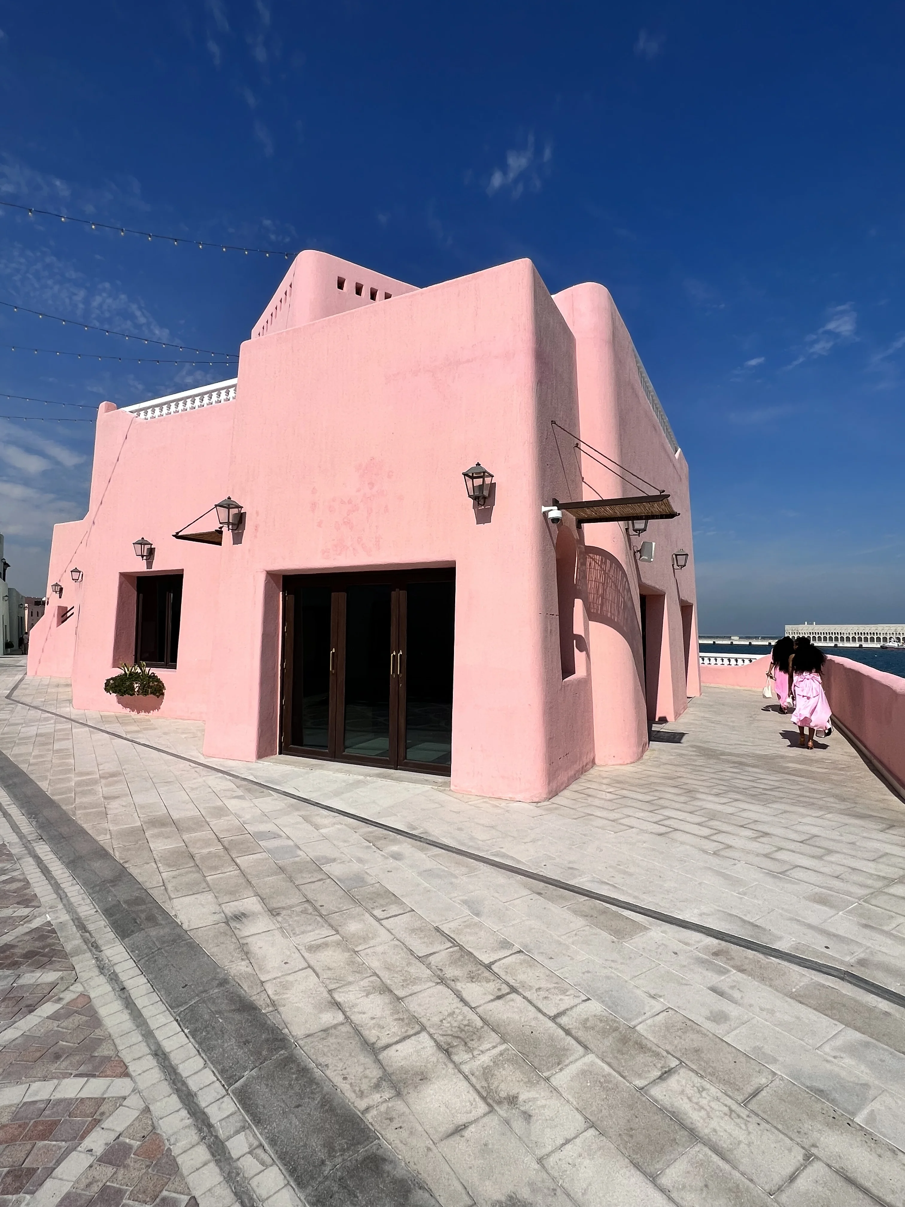 Pastel pink building along the Mina District waterfront with visitors walking past, Doha